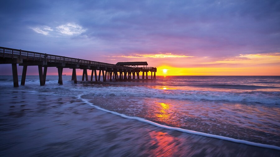 Sunrise over the ocean at Tybee Island Pier and Pavilion near Savannah, Georgia, with waves gently washing ashore and colorful reflections of orange and purple in the water