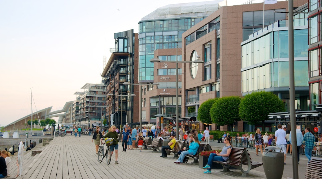 Aker Brygge que incluye arquitectura moderna y una ciudad costera y también un gran grupo de personas