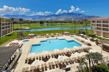 A resort pool area with multiple sun loungers and umbrellas, a central swimming pool, and a view of mountains in the background.