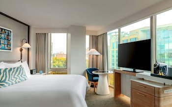 Una habitación de hotel moderna con sofá, dos mesitas auxiliares, un televisor de pantalla plana y un ventanal con vistas a la ciudad.