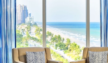 A modern living room with a large window overlooking a beach and cityscape. There are two beige armchairs with patterned cushions, a round coffee table with a vase of flowers, and a small side table.