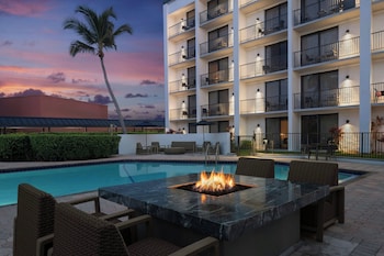 A swimming pool with lounge chairs and umbrellas, surrounded by palm trees and a building with balconies.