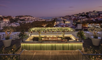 A rooftop bar with a pool table, bar area, and seating, overlooking a cityscape at dusk.