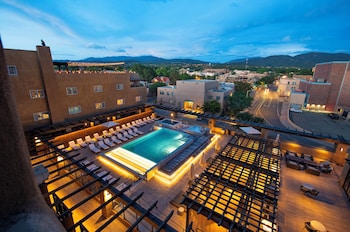 A rooftop pool area with lounge chairs and a hot tub, surrounded by buildings and a view of the mountains.