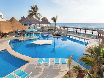 A beachfront hotel with multiple floors, a beach with blue chairs, and thatched umbrellas.