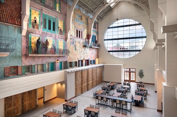 A spacious indoor atrium with multiple dining tables and chairs, a high ceiling with a mural, and a wooden wall on one side.