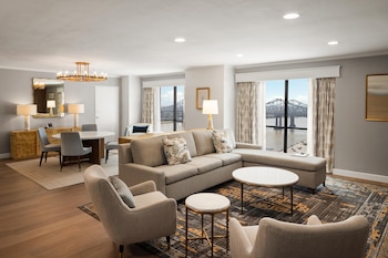 A modern hotel room with a round dining table, bar area, and large windows offering a city view.