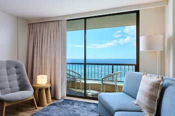 A hotel room with two beds, a desk, a chair, a TV, and a balcony with a view of the ocean and buildings.