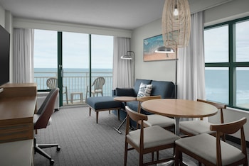 A hotel room with a balcony, a desk, a chair, a bed, and a view of the sea.