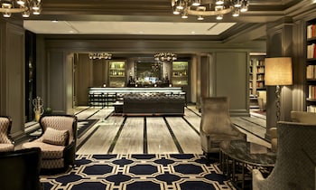 Two women walking through a hotel lobby with a large glass door, a reception desk, and a seating area with a sofa and a coffee table.