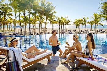 Ein Poolbereich mit Liegestühlen und Sonnenschirmen, Palmen und Blick auf den Strand.