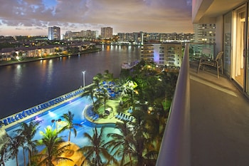 A large swimming pool surrounded by palm trees and lounge chairs, with high-rise buildings in the background.