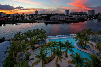 A large swimming pool surrounded by palm trees and lounge chairs, with high-rise buildings in the background.