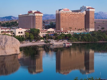 A large hotel complex with multiple buildings, a riverside promenade with sun loungers, and a boat dock.
