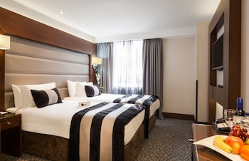 Ein Hotelzimmer mit einem großen Bett, einem Nachttisch mit Telefon, einem Sessel, einem Tisch mit Champagnerflasche und einer Vase mit Blumen.