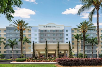 A hotel named Homewood Suites by Hilton with a reflection in a calm water body, surrounded by trees and well-maintained landscaping.