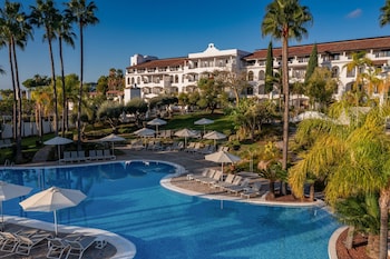 A resort with a large swimming pool, lounge chairs, and umbrellas. There are palm trees and a view of a golf course and mountains in the background.