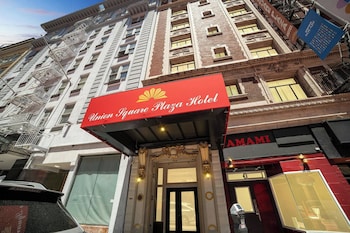 L’entrée d’un hôtel, avec un auvent rouge, une grande porte vitrée et une enseigne indiquant « Hotel Squared Plaza Hotel ».