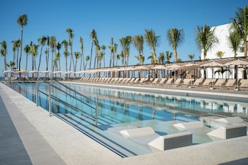 Zona de piscina con sillones de descanso y sombrillas, rodeada de palmeras.