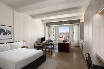 Habitación de hotel moderna con una cama grande, un escritorio, una silla, una mesita, una lámpara y vistas de la ciudad a través de amplias ventanas.