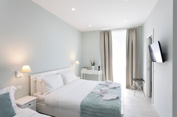 A hotel room with a neatly made bed, a bedside table with a lamp, a small desk with a chair, a TV mounted on the wall, and a window with curtains.