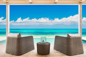 Un sofá de mimbre con cojines y un candelabro de vidrio frente a una piscina, con vista al mar turquesa y un cielo despejado.