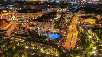 An aerial view of a cityscape at night with illuminated buildings, roads, and a large central plaza with a fountain.