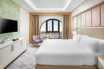 Een moderne hotelkamer met een groot rondboogvenster, een flatscreen televisie aan de muur, een groene fauteuil, een beige bank en een ronde bijzettafel met een lamp.