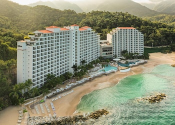 Un balcón con sillas de mimbre y cojines rojos, con vista a una playa con formaciones rocosas y agua azul cristalina.