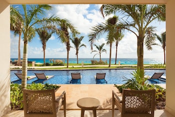A resort pool with two people swimming, surrounded by palm trees and a view of the ocean.