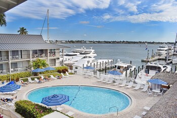 A covered outdoor seating area with wicker furniture, a table with a newspaper and a mug, and a view of boats at a marina.