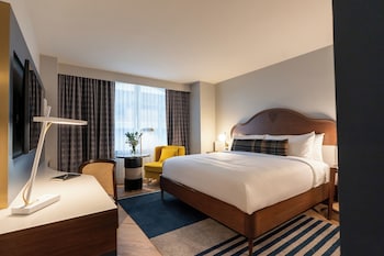 A hotel room with a large bed, a desk with a lamp, a chair, a TV, and a window with curtains.