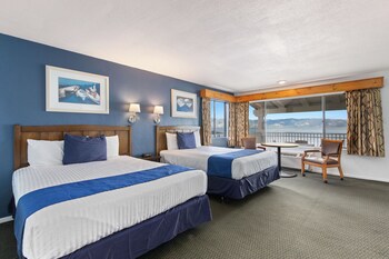 Habitación de hotel con una cama grande, dos sillas, una mesa y vistas al mar y las montañas a través de amplias ventanas.