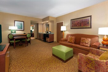 A hotel room with a dining table, green chairs, a sofa, a TV, a kitchen area, and a large framed picture on the wall.