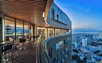 A high-rise hotel with a modern design, featuring a rooftop bar with tables and chairs, large windows, and a view of the cityscape.