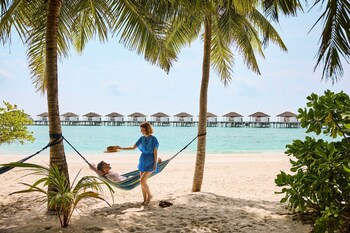 Vue aérienne d’un complexe hôtelier avec des bungalows sur pilotis reliés par une passerelle, entouré d’eaux turquoise et d’une plage de sable blanc.