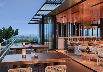 Un bar moderne situé sur un toit-terrasse, avec des tables en bois et des chaises en osier, de grandes fenêtres offrant une vue sur la ville et un éclairage d’ambiance.