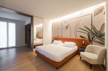 A bedroom with a large bed, a desk with a chair, a TV mounted on the wall, and a window with a view of the outside.