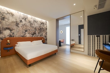 A bedroom with a large bed, a desk with a chair, a TV mounted on the wall, and a window with a view of the outside.