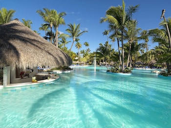 A resort pool with clear water, surrounded by palm trees and thatched-roof structures.
