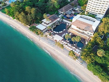 A beachside lounge area with white cushioned seating and umbrellas, overlooking a clear blue sea and palm trees.