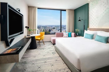 Ein Zimmer mit großem Fenster und Blick auf die Stadt, ein Bett mit Frühstücksbretzel und ein gelber Sessel mit Kissen.