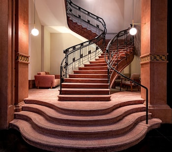 A corridor with a polished floor, flanked by potted plants and chairs, leading to a central area with a decorative ceiling and a door at the end.