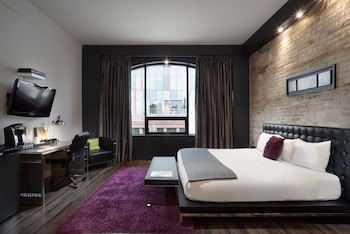 A modern hotel room with a brick wall, two beds, a desk, and a large window.