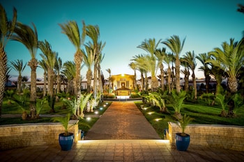 A pathway leading to a building with a central entrance, flanked by palm trees and illuminated lights.