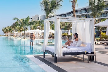 A resort pool with clear blue water, surrounded by palm trees and lounge chairs, with a view of the ocean and modern buildings in the background.