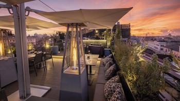 An outdoor terrace with tables and chairs, a large umbrella, and a city view at sunset.