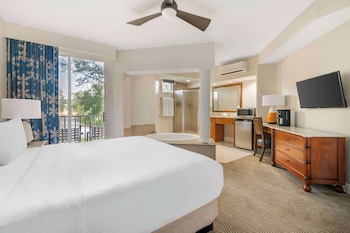 A hotel room with a large bed, a desk with a flat-screen TV, a bathroom with a sink and mirror, and a balcony with blue curtains.