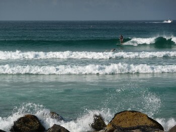 Una playa con olas, una persona practicando surf y rocas al frente.