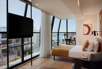 Una habitación de hotel moderna con una cama grande, un escritorio y una televisión. La habitación tiene ventanas amplias con vistas a la ciudad.
